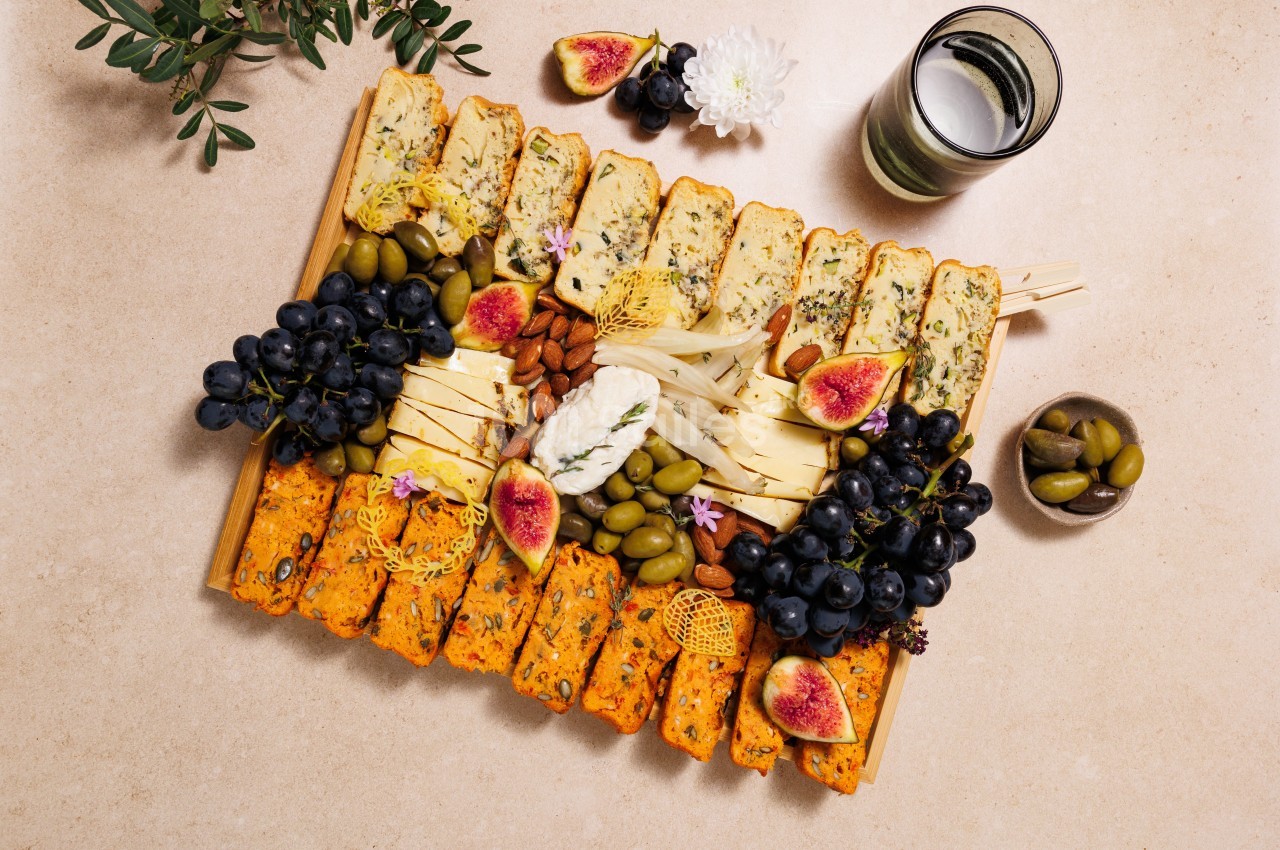 Plateau garni de fromages, fruits frais, olives, noix et tranches de pain, disposé sur une table beige.