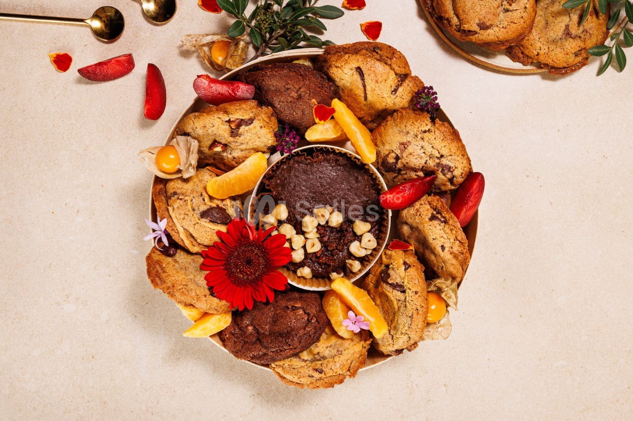 Assortiment de cookies et de fruits disposés autour d'un gâteau au chocolat, décoré de fleurs et de noix.