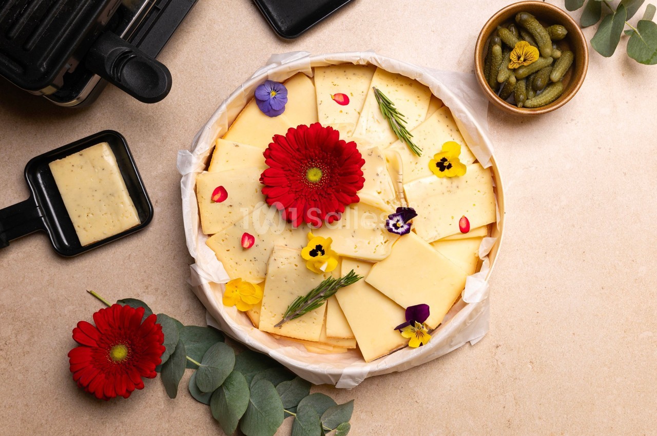 Plateau de fromage décoré de fleurs comestibles, accompagné de cornichons et d'un appareil à raclette.