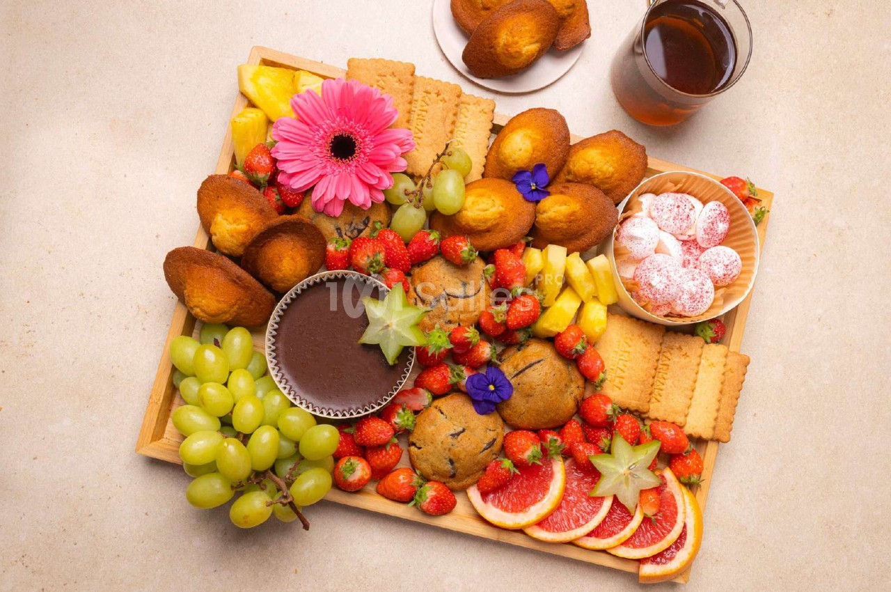 Plateau garni de fruits, biscuits, madeleines, muffins, chocolat fondu et une boisson sur une table beige.