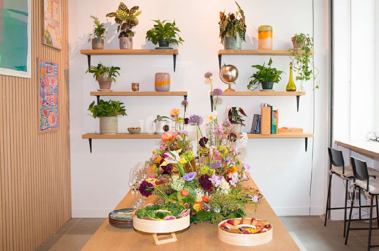Table en bois décorée de fleurs colorées, entourée d'étagères avec des plantes et objets dans un espace lumineux.