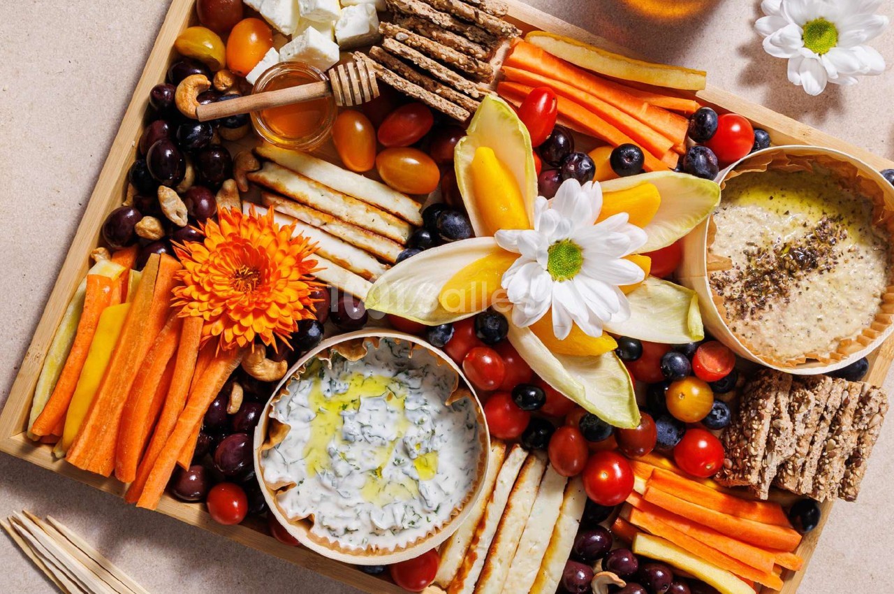 Plateau coloré de crudités, fromages, dips, fruits secs et fleurs comestibles disposés de manière artistique.