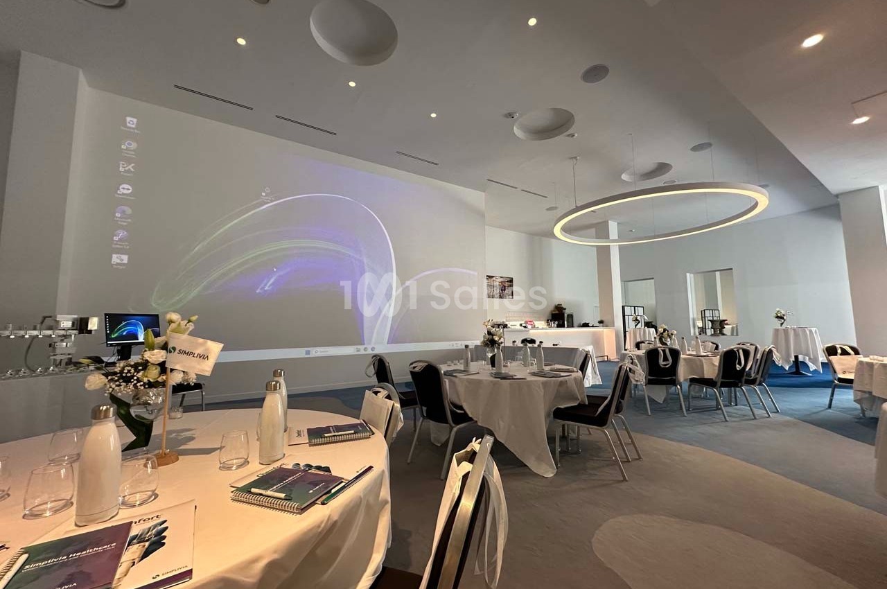 Salle de conférence lumineuse avec tables rondes dressées, projecteur allumé et décor moderne.