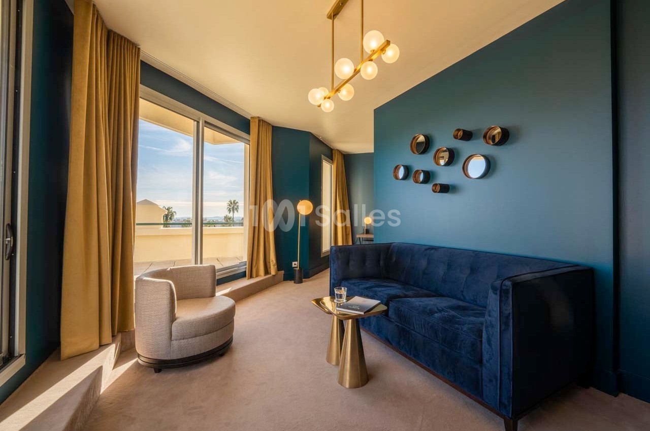 Salon moderne avec canapé bleu, fauteuil beige, table dorée et grandes fenêtres offrant une vue sur l'extérieur.