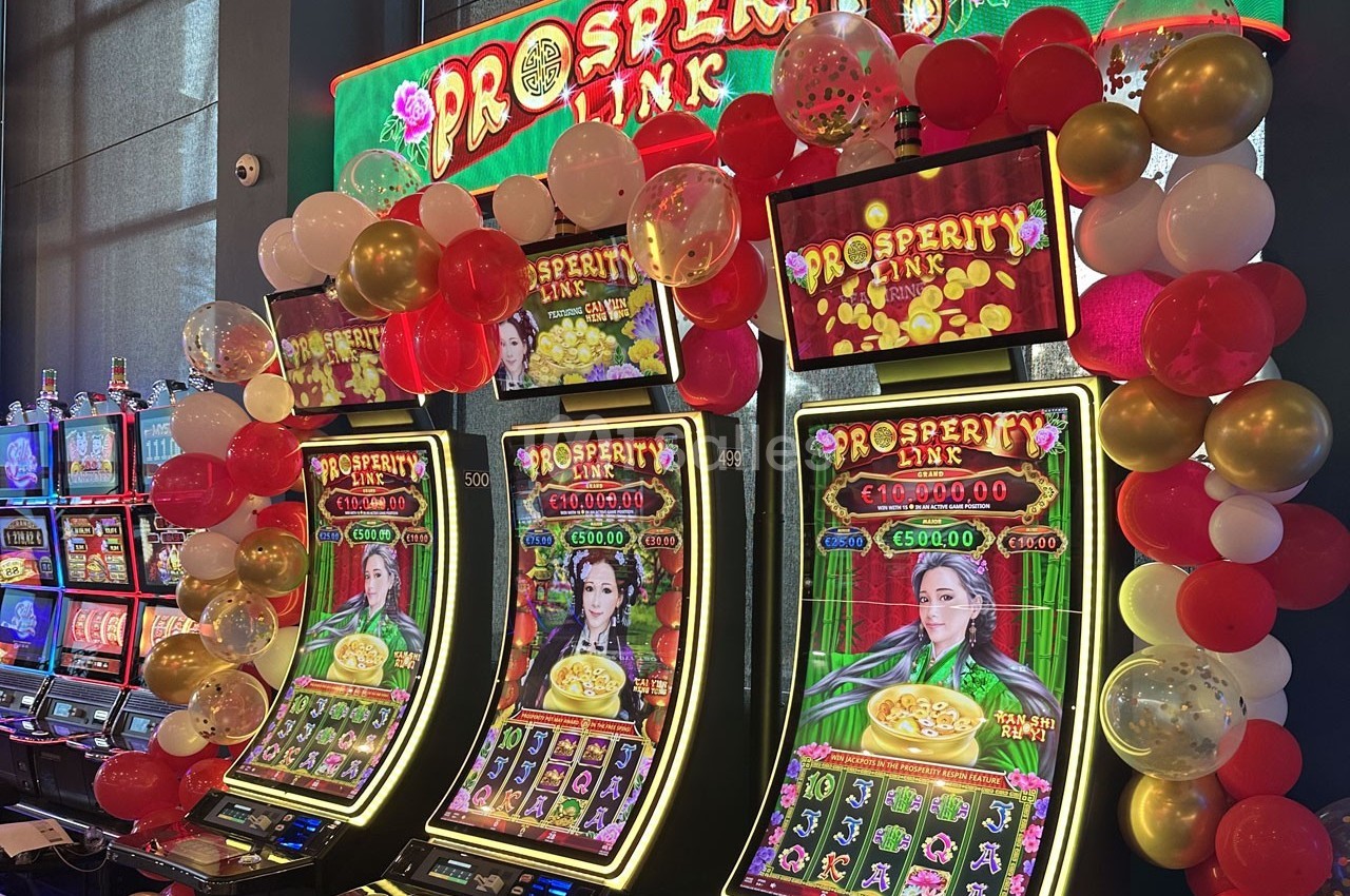 Machines à sous lumineuses décorées de ballons, affichant le jeu ’Prosperity Link’ dans un casino.