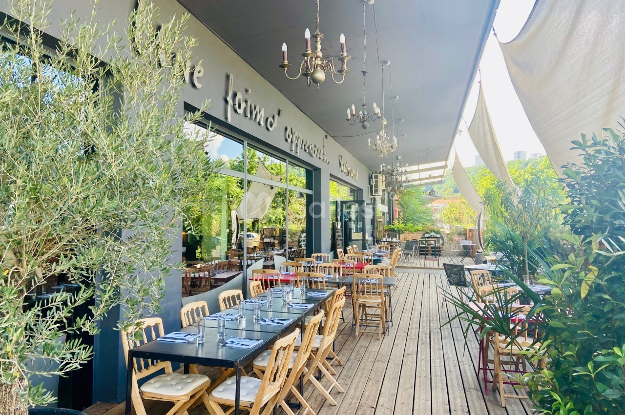 Terrasse d'un restaurant avec tables en bois, chaises pliantes et végétation, sous un auvent avec des luminaires suspendus.