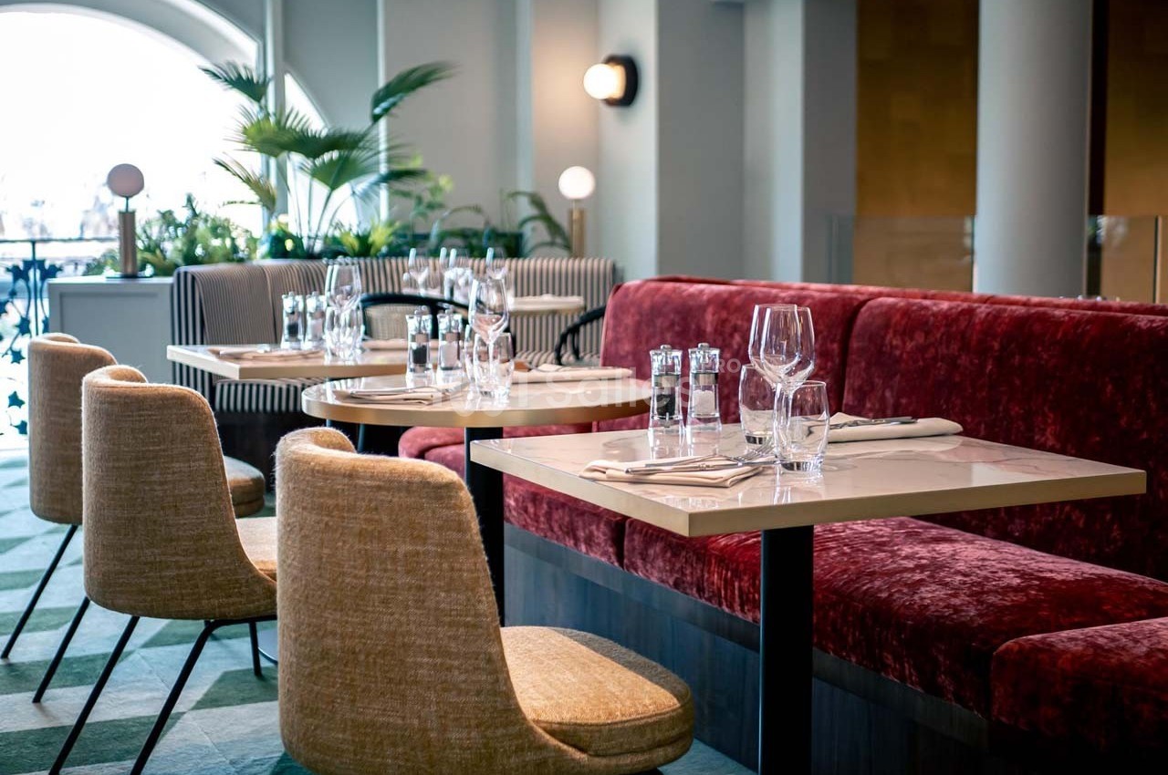Salle de restaurant lumineuse avec tables dressées, chaises rembourrées et banquettes rouges, décor moderne et végétation.