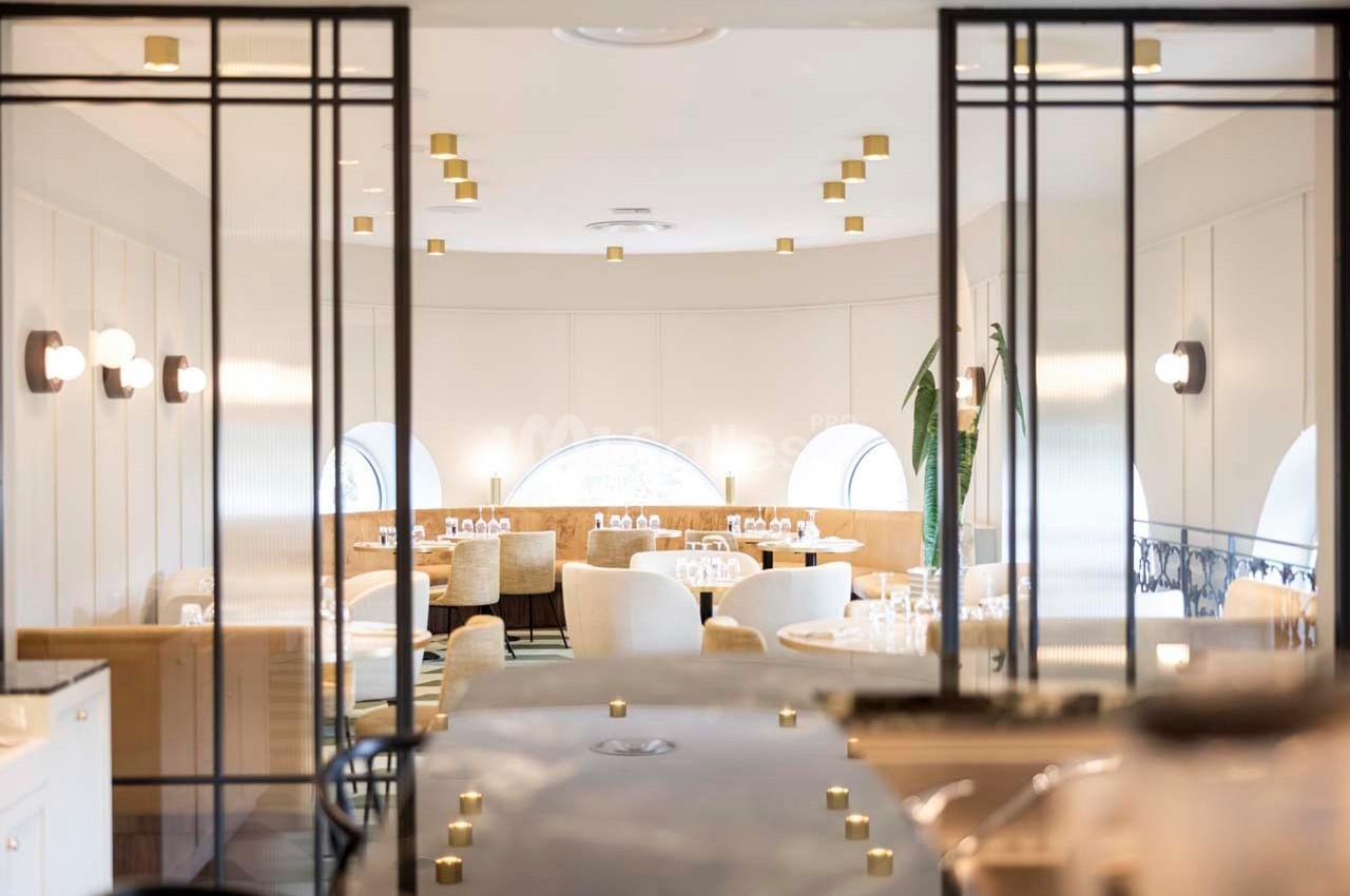 Salle de restaurant lumineuse avec tables dressées, chaises rembourrées et grandes fenêtres en arrière-plan.