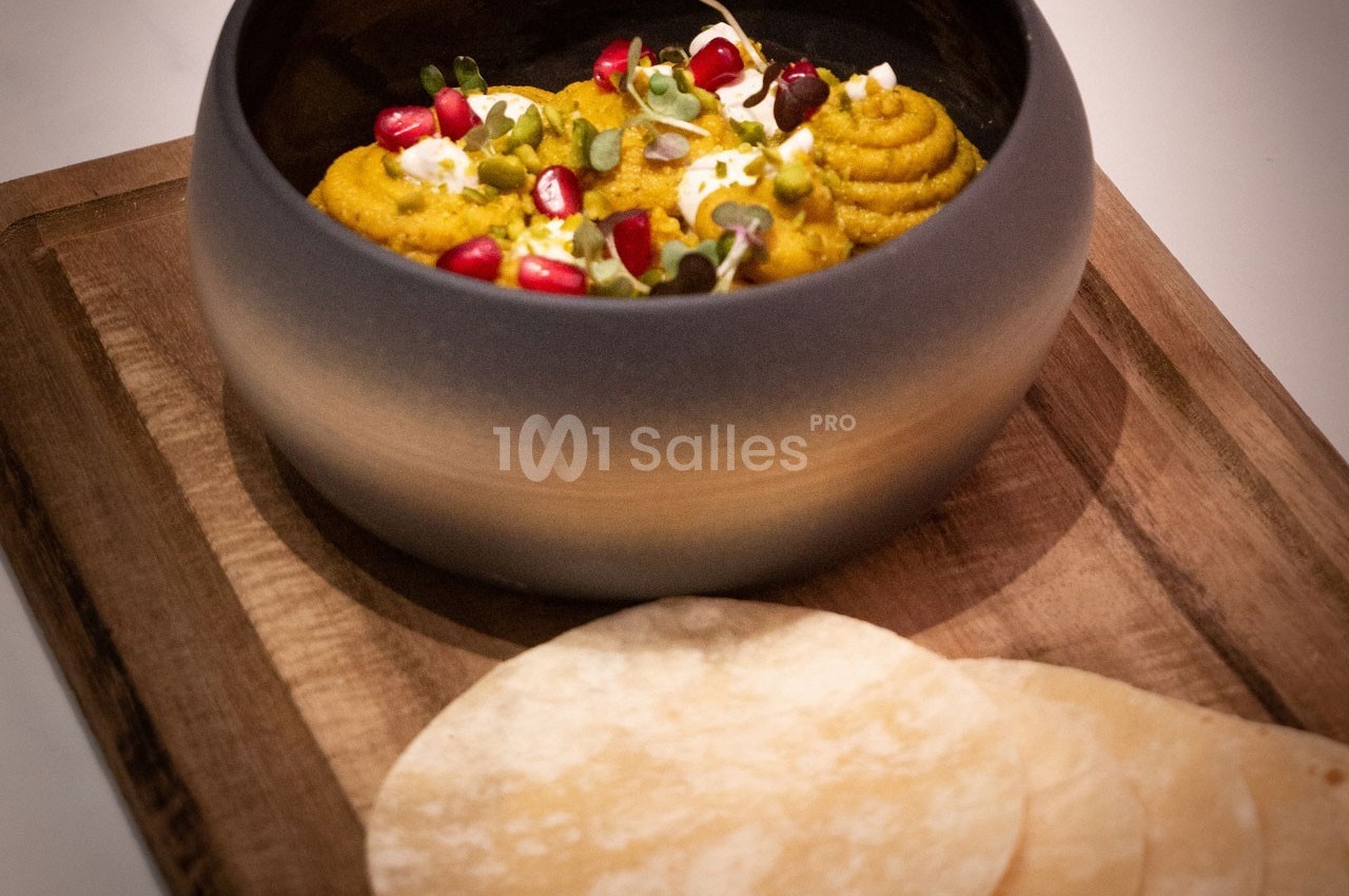 Bol de purée garnie de graines de grenade et herbes fraîches, accompagné de galettes sur une planche en bois.