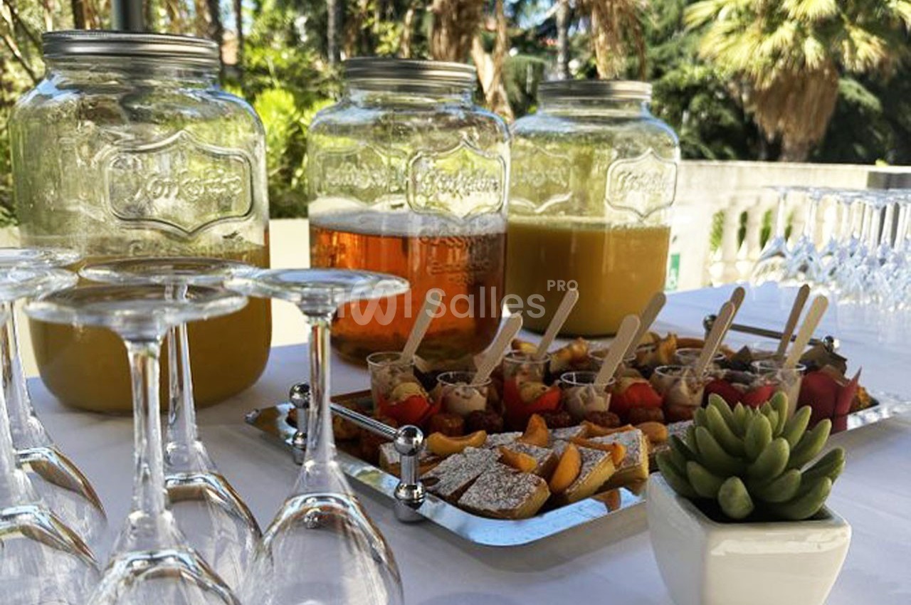 Buffet extérieur avec boissons en jarres, verrines sucrées et salées, verres vides et plante décorative.