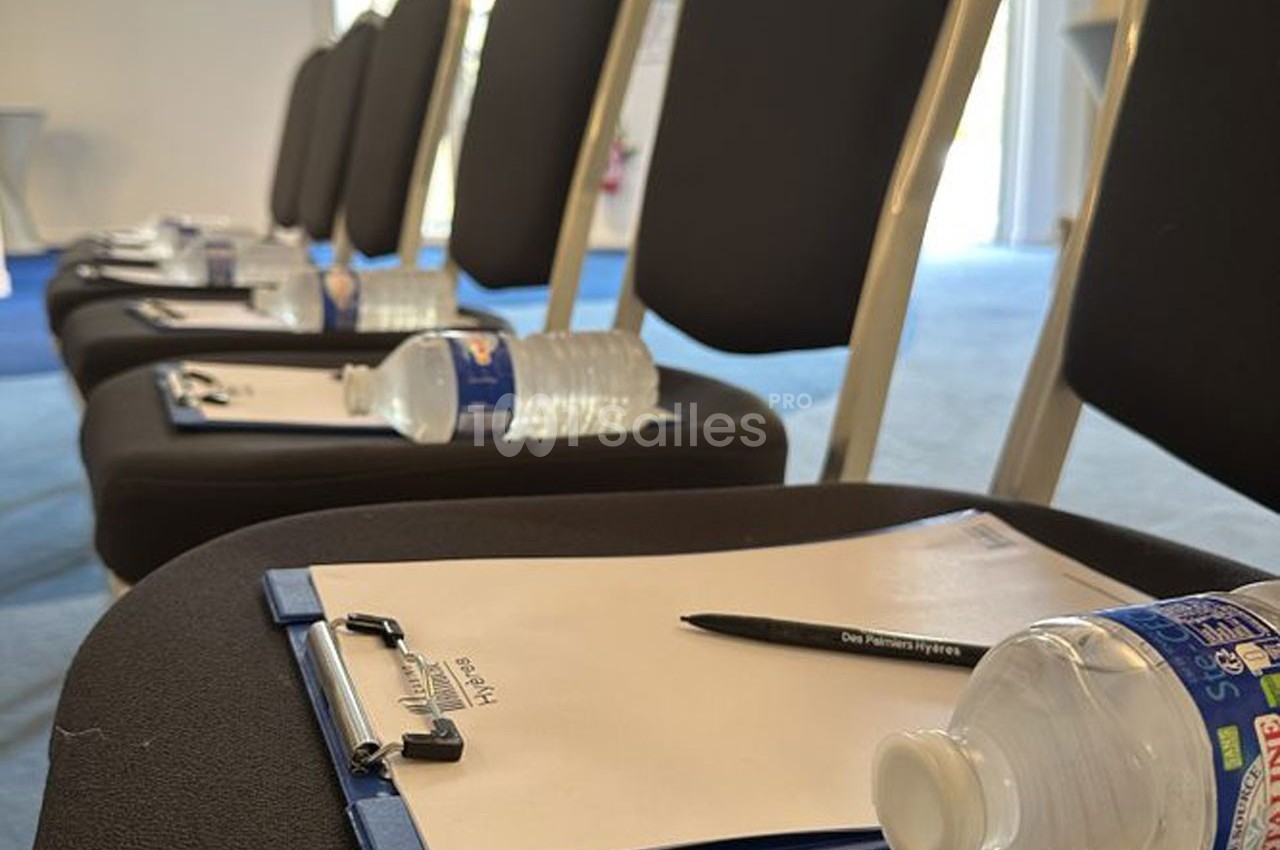 Chaises alignées avec des blocs-notes, stylos et bouteilles d'eau posés sur chaque siège dans une salle de réunion.