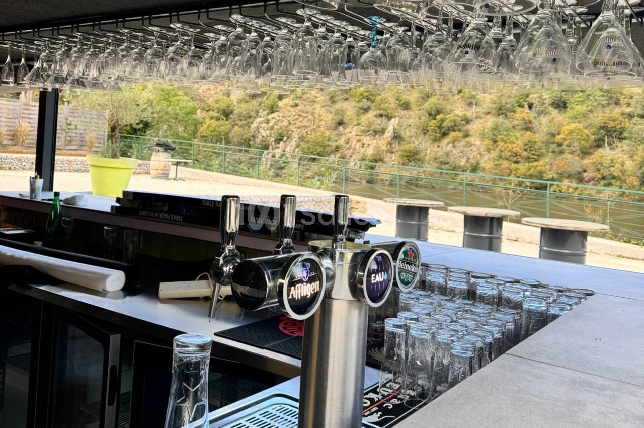 Comptoir de bar extérieur avec tireuses à bière, verres suspendus et vue sur un paysage naturel en arrière-plan.