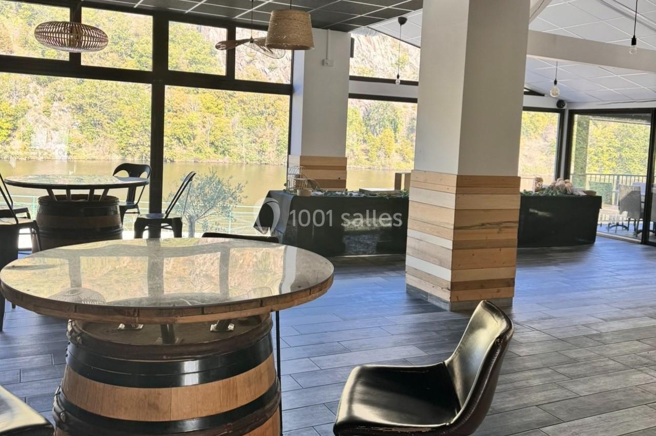 Salle lumineuse avec grandes baies vitrées, tables en tonneaux, chaises noires et vue sur un paysage naturel verdoyant.