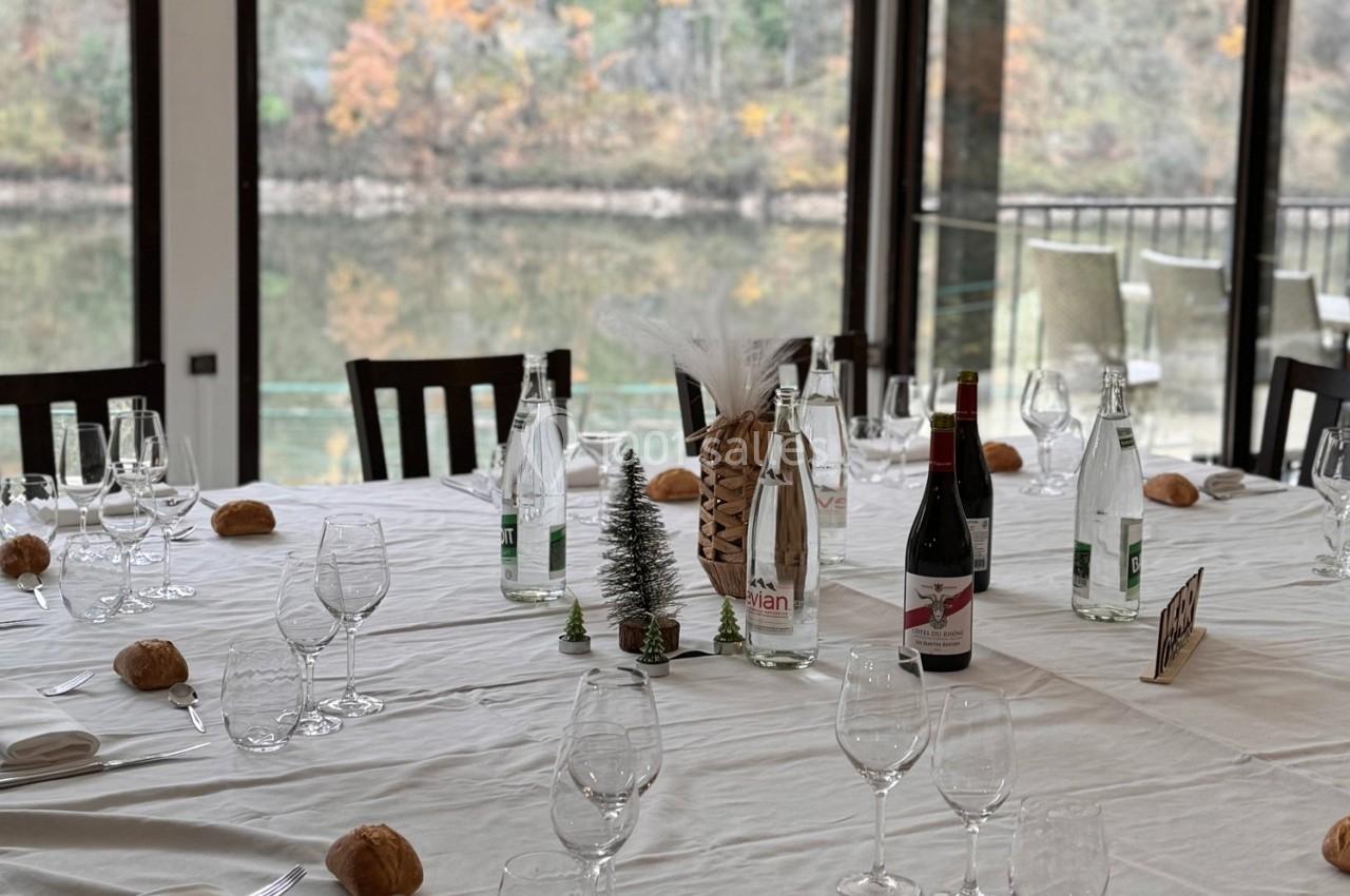 Table dressée avec nappes blanches, verres, bouteilles d'eau et de vin, petits pains, décorations et vue sur un lac.