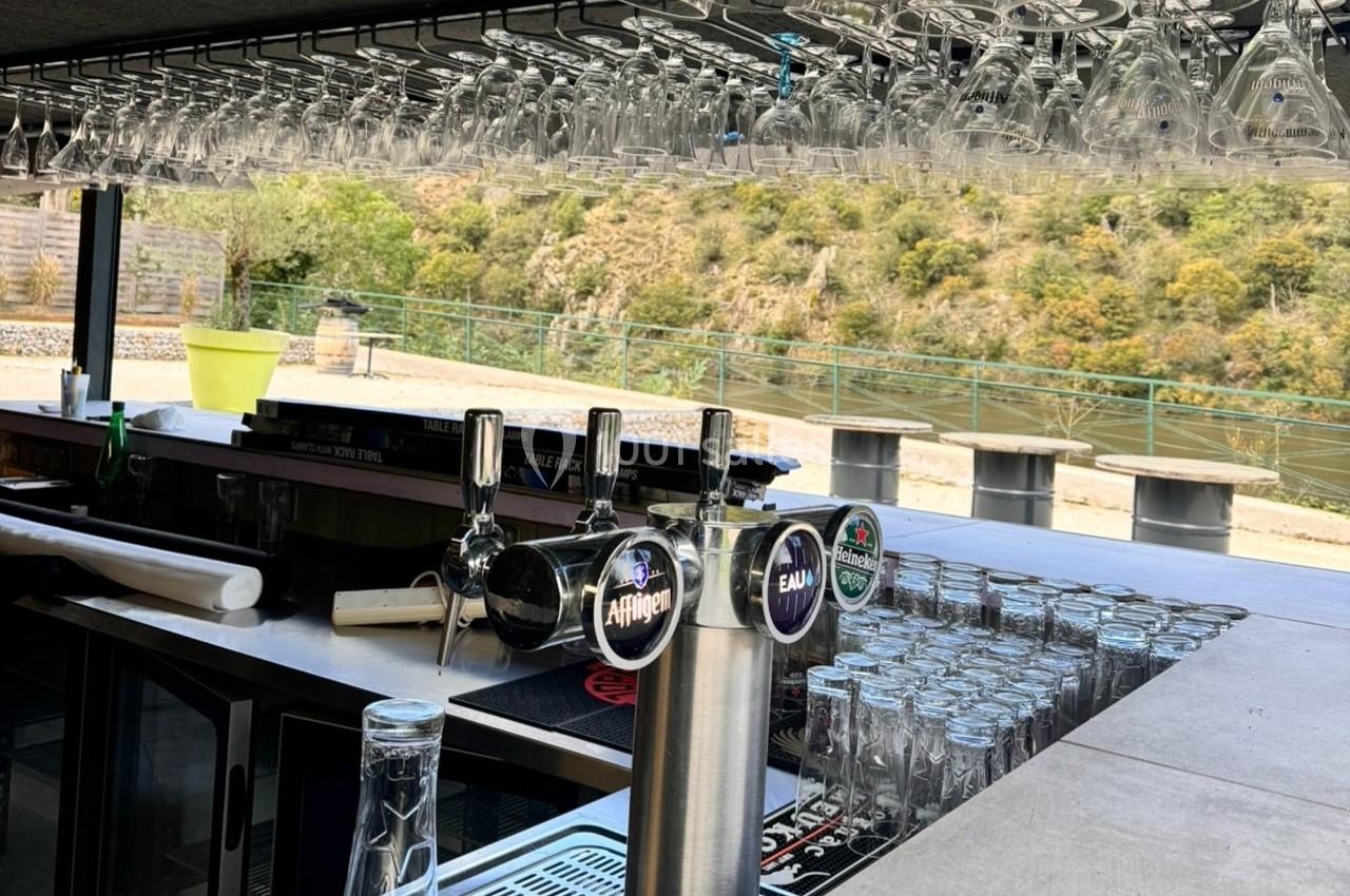 Comptoir de bar extérieur avec tireuses à bière, verres suspendus et vue sur un paysage naturel en arrière-plan.