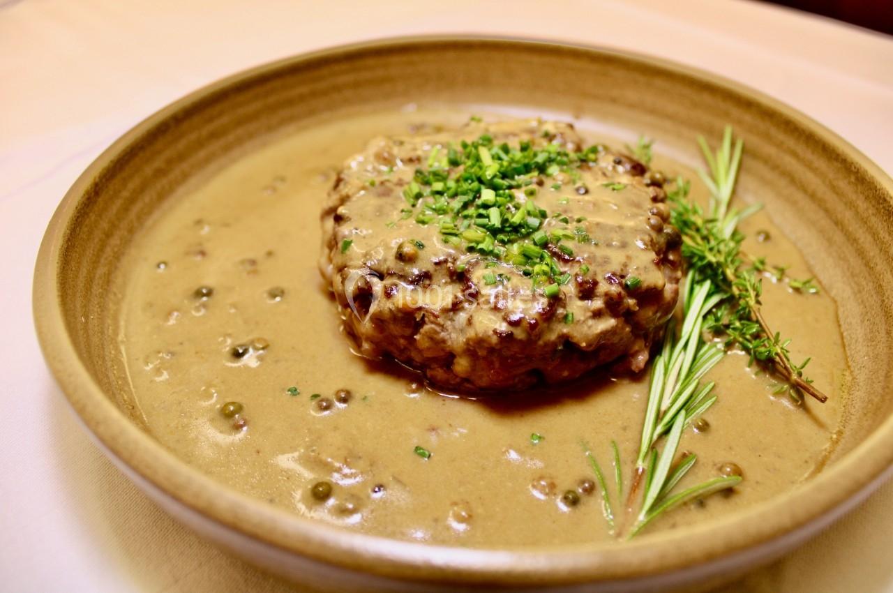 Plat de steak haché nappé de sauce au poivre, garni de ciboulette, romarin et thym, servi dans une assiette beige.