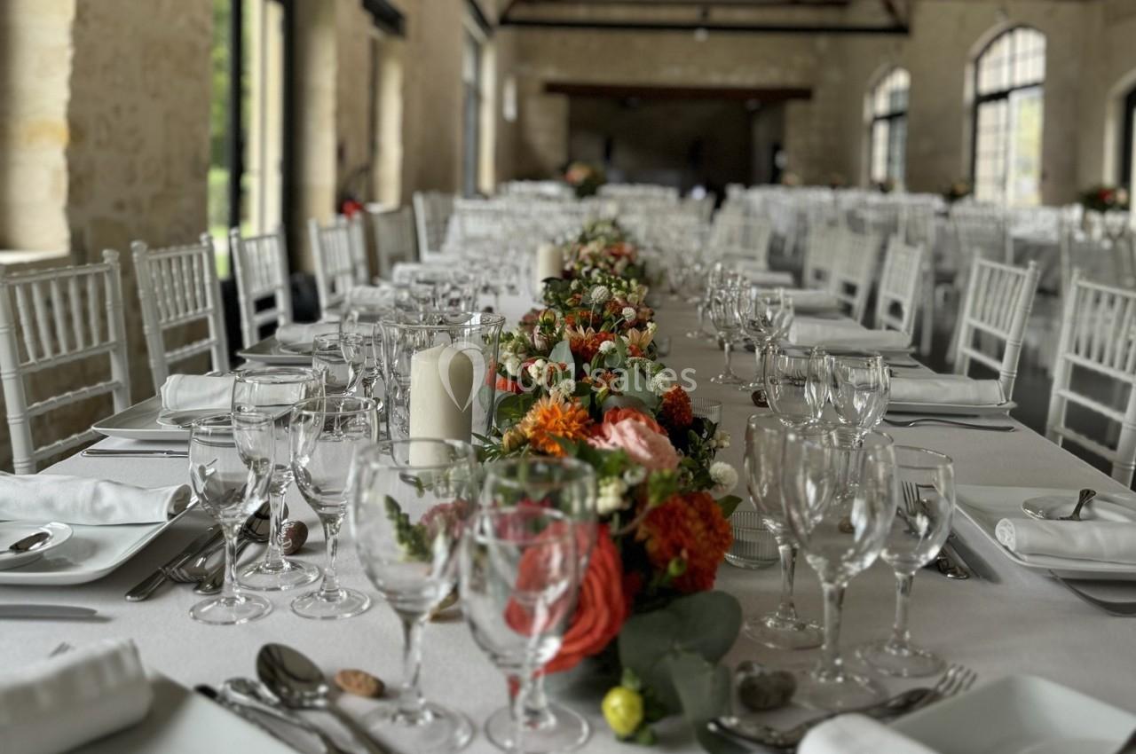 Grande table de banquet décorée de fleurs colorées, bougies blanches et vaisselle élégante dans une salle en pierre.