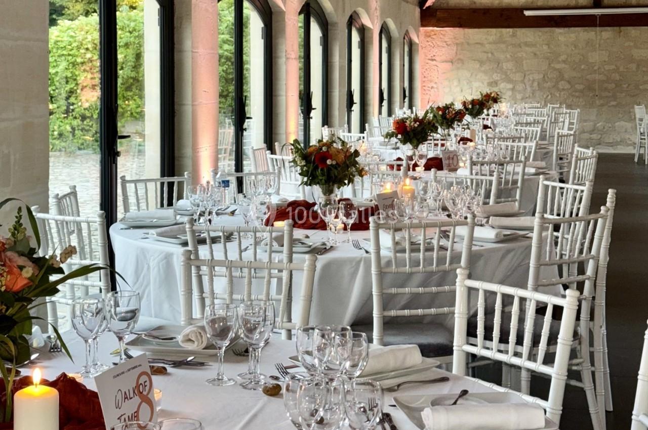 Salle de réception lumineuse avec tables dressées, nappes blanches, chaises blanches et décorations florales.