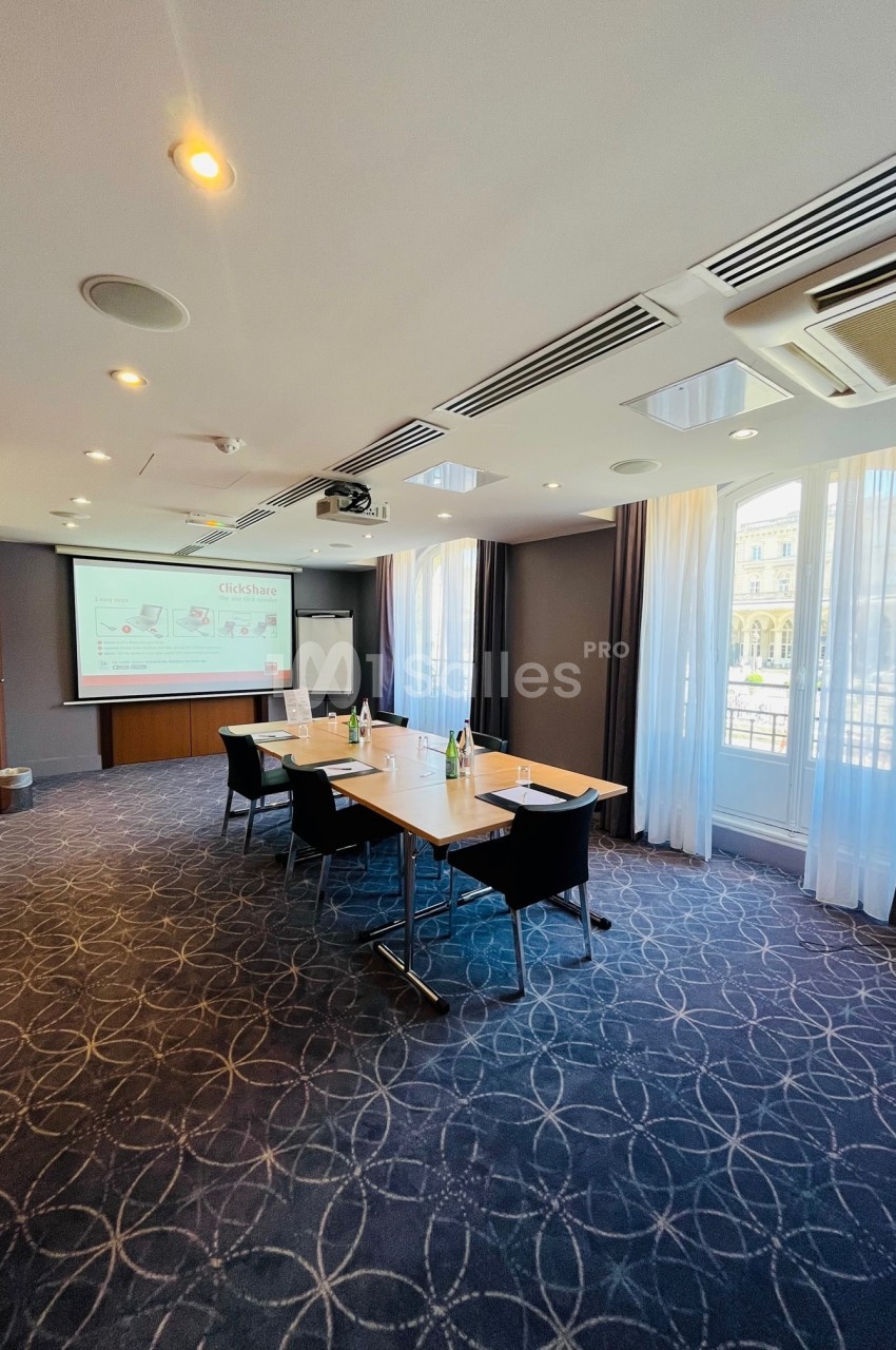 Salle de réunion lumineuse avec table rectangulaire, chaises noires, écran de projection et grandes fenêtres.