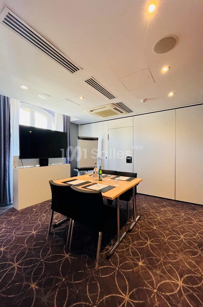 Salle de réunion lumineuse avec table, chaises, écran, paperboard et rideaux encadrant une fenêtre.