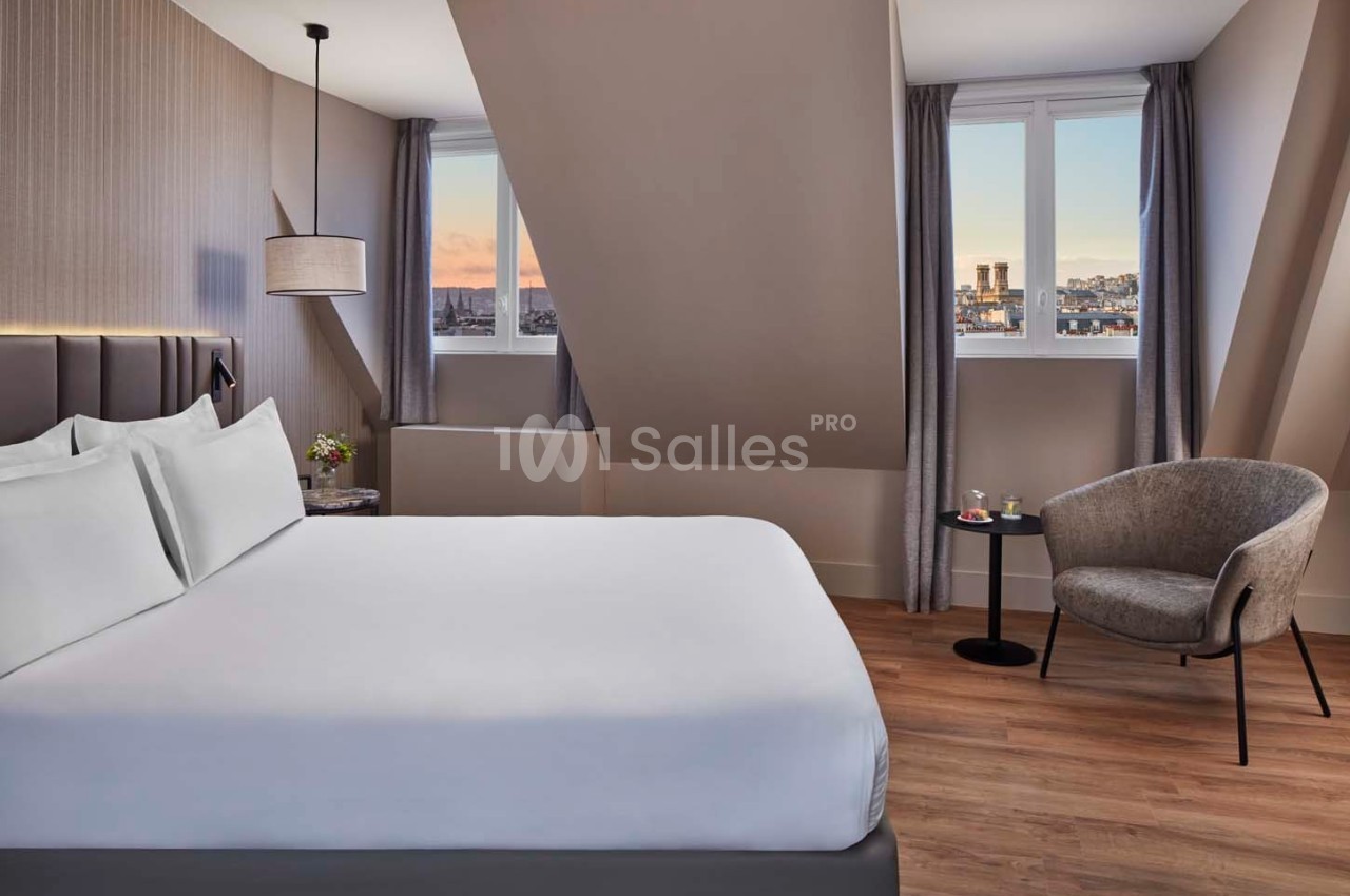 Chambre moderne avec lit double, fauteuil, table basse et vue sur la ville à travers deux fenêtres mansardées.