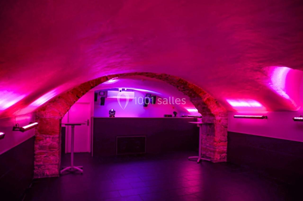 Salle voûtée en pierre avec éclairage rose et violet, murs blancs et sol sombre, équipée de tables hautes.