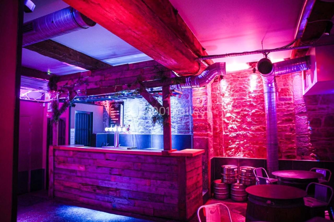 Bar en bois avec éclairage violet et rose, tonneaux en métal au sol et murs en pierre apparente.