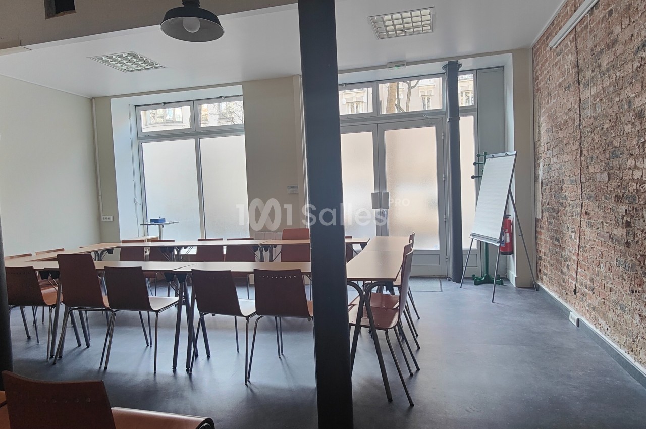 Salle de réunion lumineuse avec des tables disposées en U, des chaises, un tableau blanc et un mur en briques apparentes.