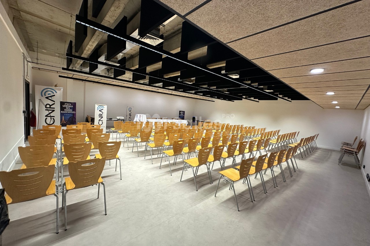 Salle de conférence vide avec rangées de chaises en bois alignées, plafond avec poutres noires et éclairage intégré.