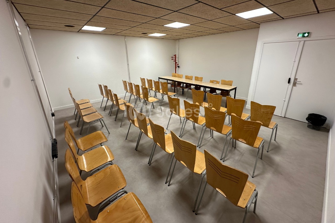 Salle de réunion avec des rangées de chaises en bois disposées face à une table et un tableau blanc.