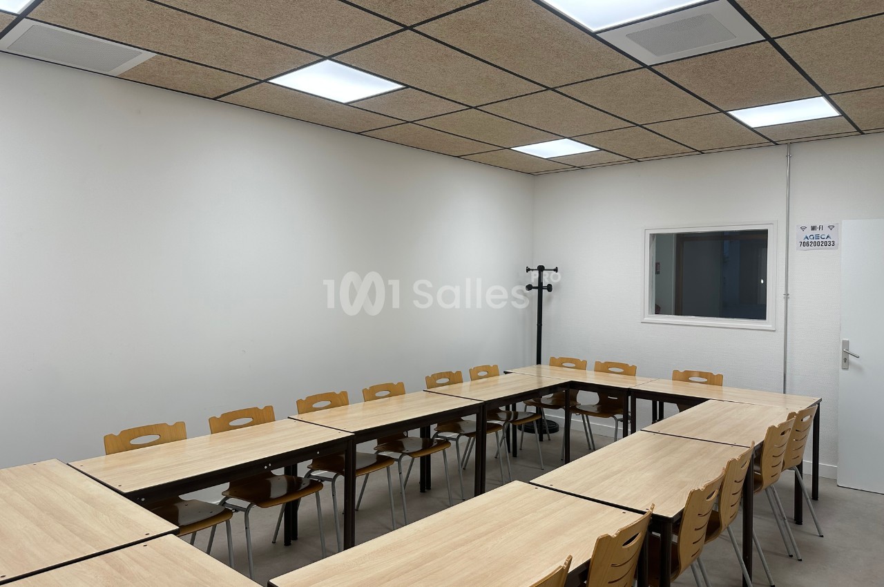 Salle de réunion avec des tables disposées en U, des chaises en bois et un mur blanc éclairé par des plafonniers.