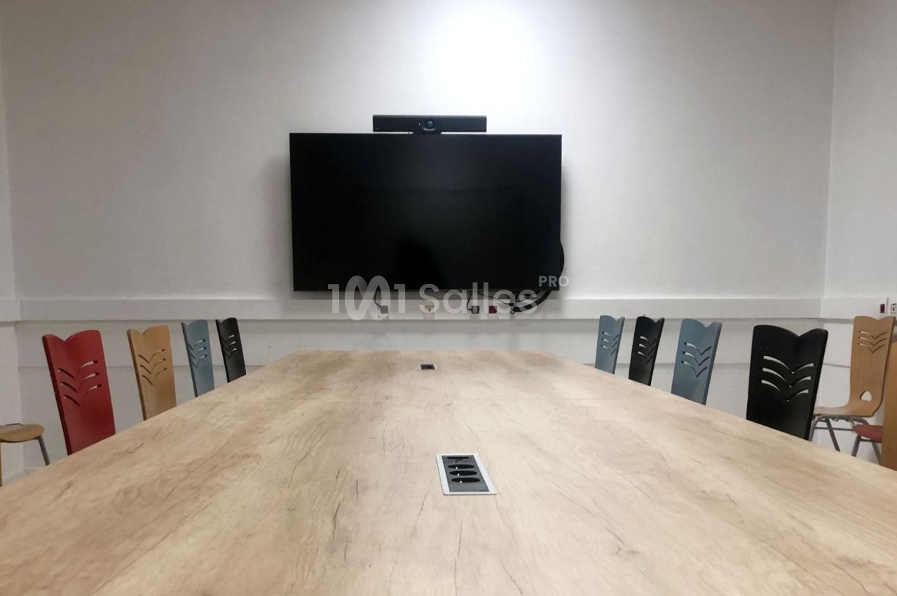 Salle de réunion avec une grande table en bois, des chaises colorées et un écran mural noir.