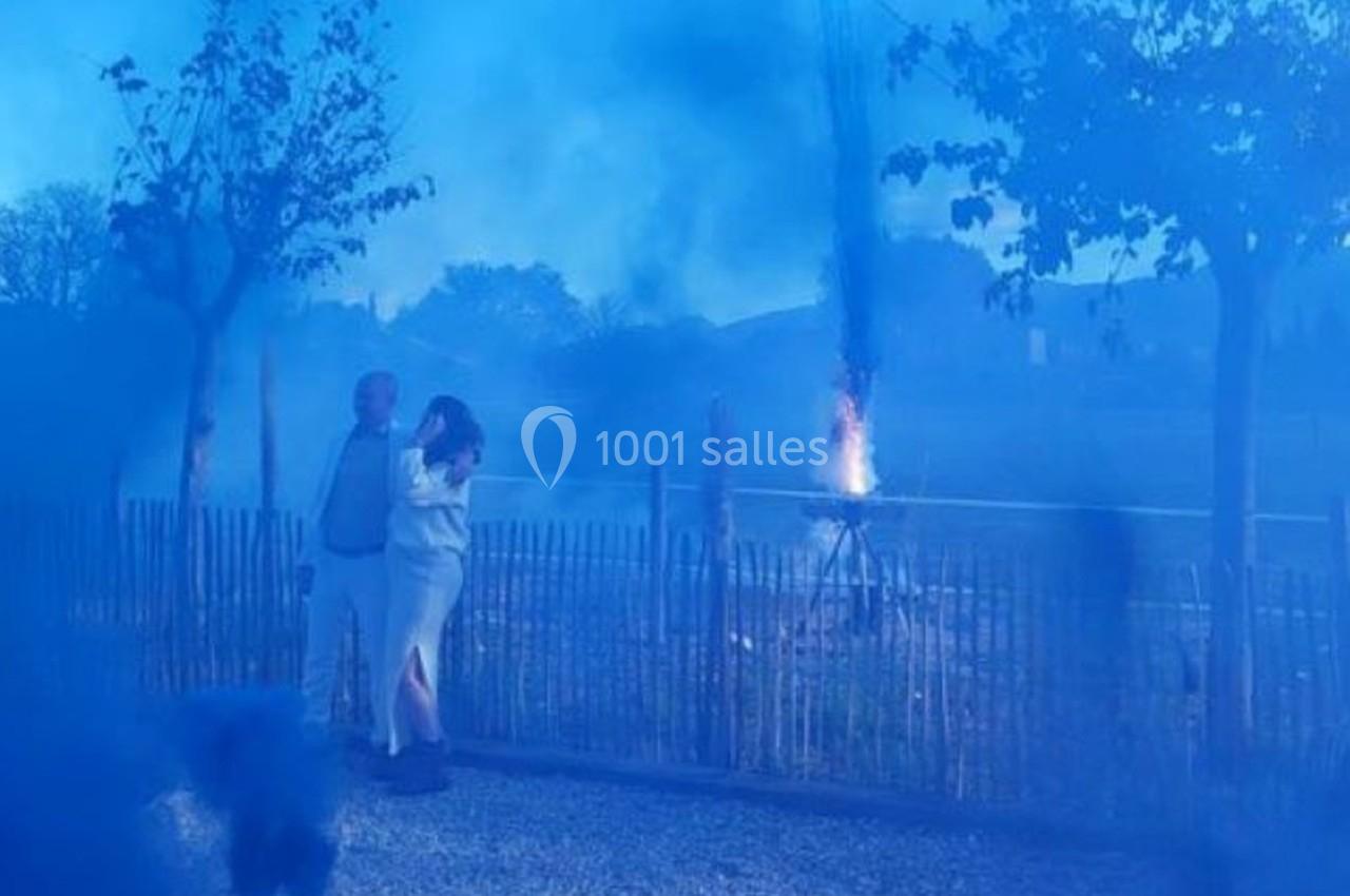 Un couple debout près d'une clôture observe un feu allumé à distance, enveloppé dans une épaisse fumée bleue.