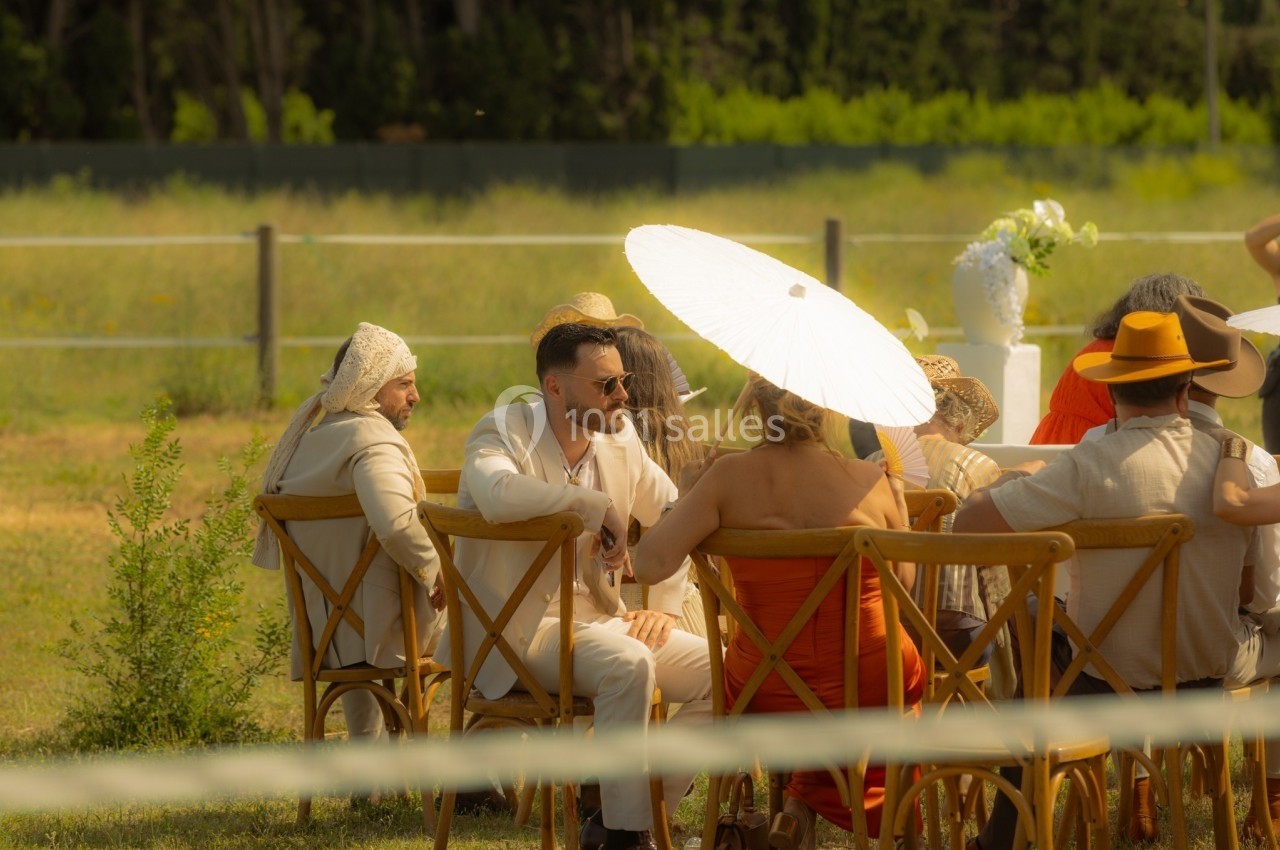 Groupe de personnes assises sur des chaises en bois lors d'un événement en extérieur, sous un parasol blanc.