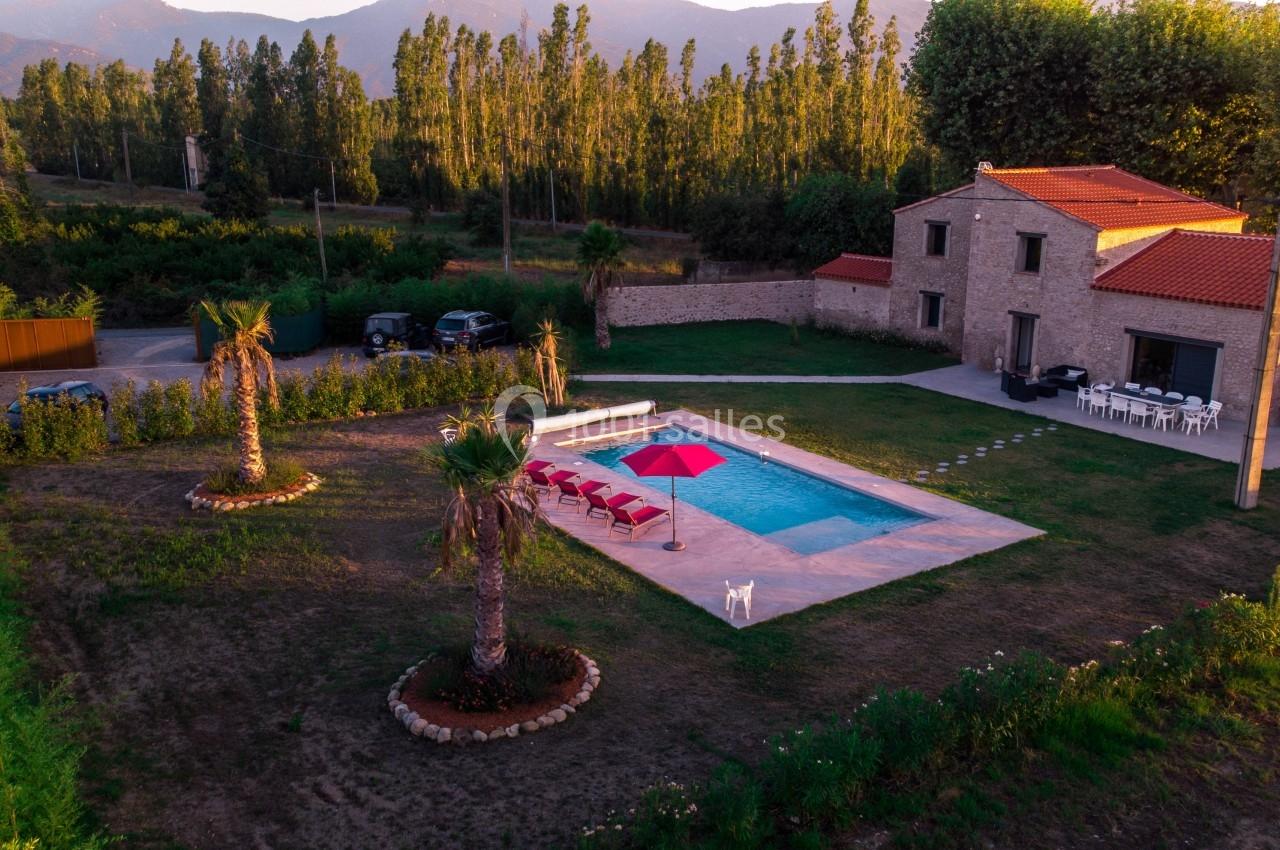 Vue aérienne d'une maison en pierre avec piscine, transats rouges et parasol, entourée de verdure et de montagnes.