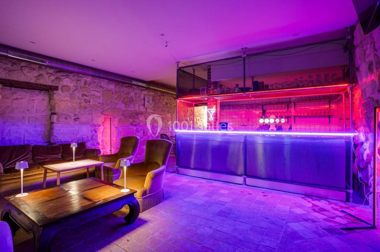 Bar avec comptoir éclairé par des néons violets, fauteuils et tables en bois dans une ambiance de pierre apparente.