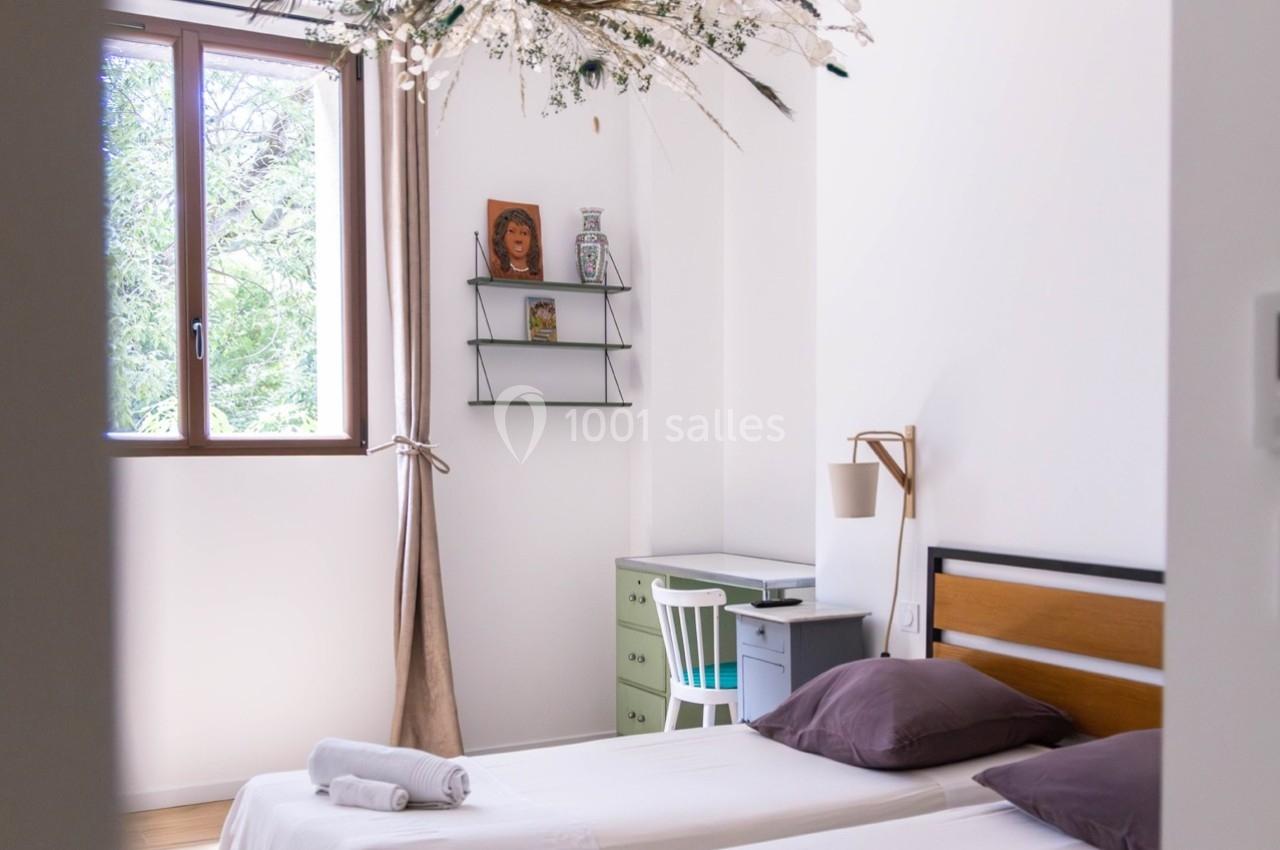 Chambre lumineuse avec deux lits simples, un bureau, une étagère murale et une fenêtre donnant sur des arbres.
