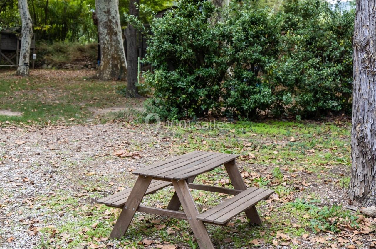 Table de pique-nique en bois installée sur un sol parsemé de feuilles, entourée d'arbres et de buissons.