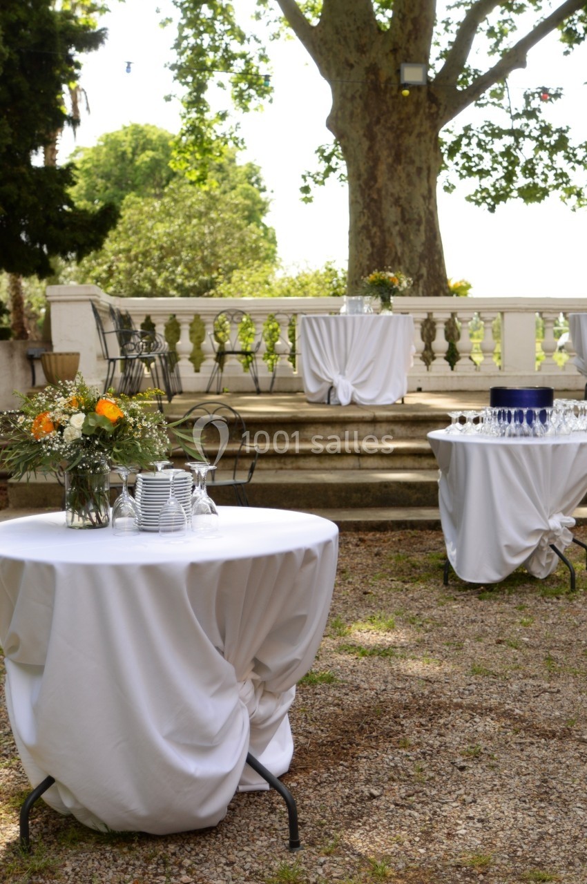 Tables rondes drapées de nappes blanches avec vaisselle et fleurs, disposées en extérieur près d'escaliers en pierre.
