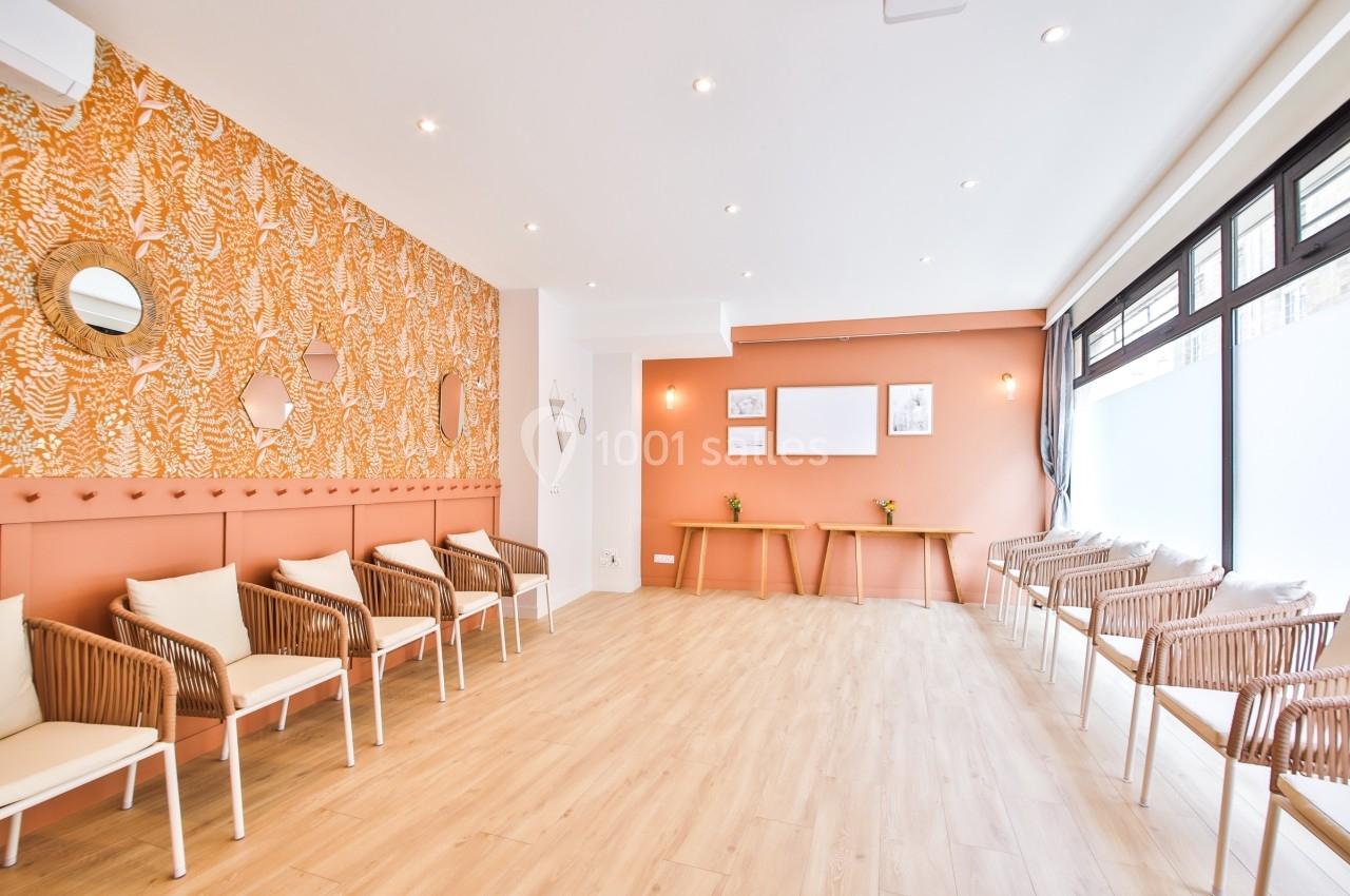 Salle lumineuse avec murs orange, parquet clair, chaises en rotin et grandes fenêtres laissant entrer la lumière naturelle.