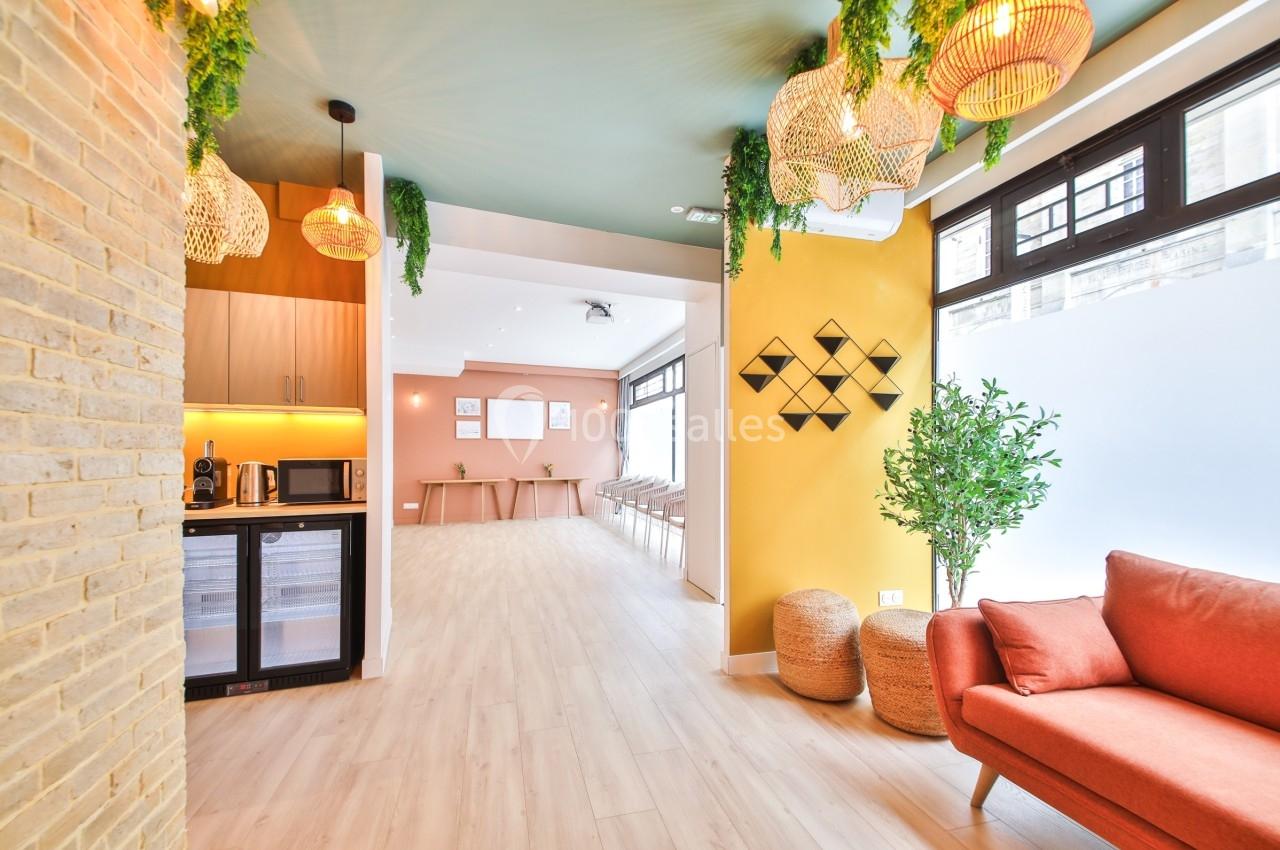 Espace intérieur lumineux avec canapé orange, plantes, luminaires suspendus et cuisine équipée en arrière-plan.