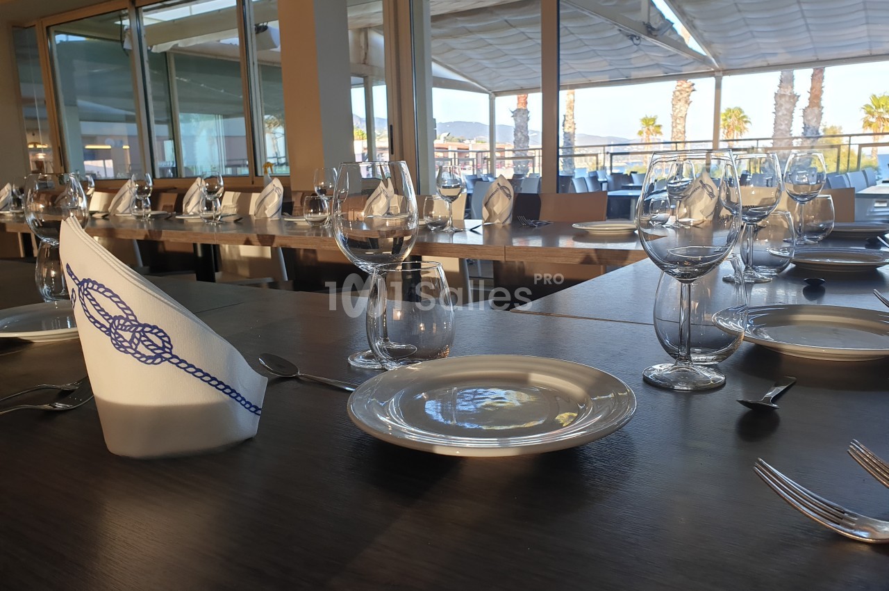 Table dressée avec assiettes, verres et serviettes pliées, dans un restaurant avec vue sur une terrasse ensoleillée.