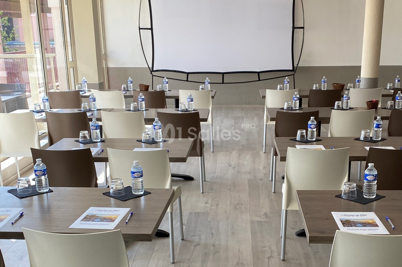 Salle de réunion lumineuse avec des tables disposées en rangées, équipées de bouteilles d'eau, stylos et documents.