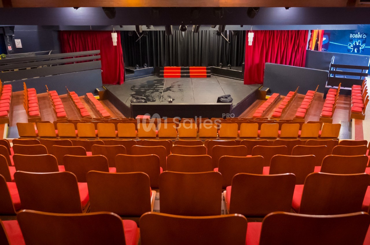 Salle de théâtre vide avec des rangées de sièges rouges face à une scène entourée de rideaux rouges.