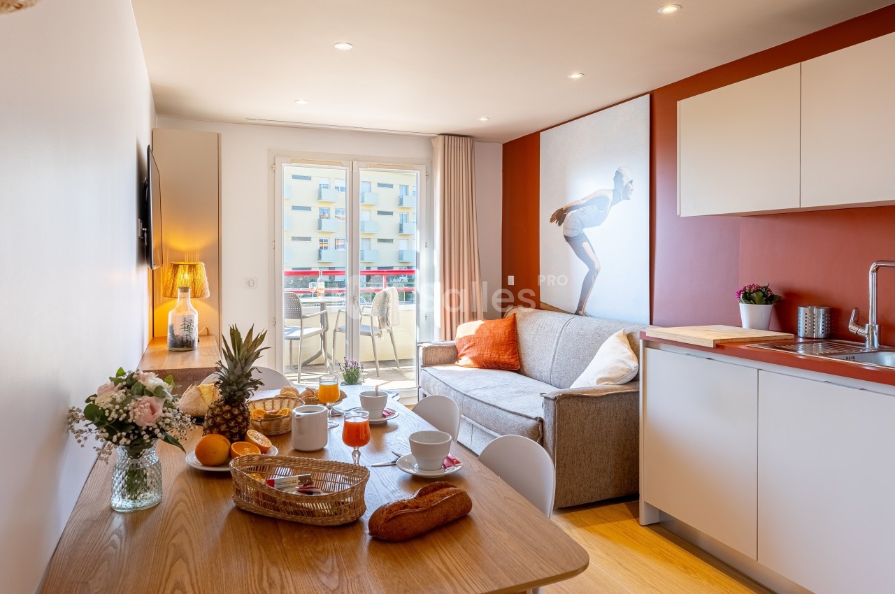 Salon avec kitchenette, table dressée pour le petit-déjeuner, canapé, et balcon donnant sur des immeubles.