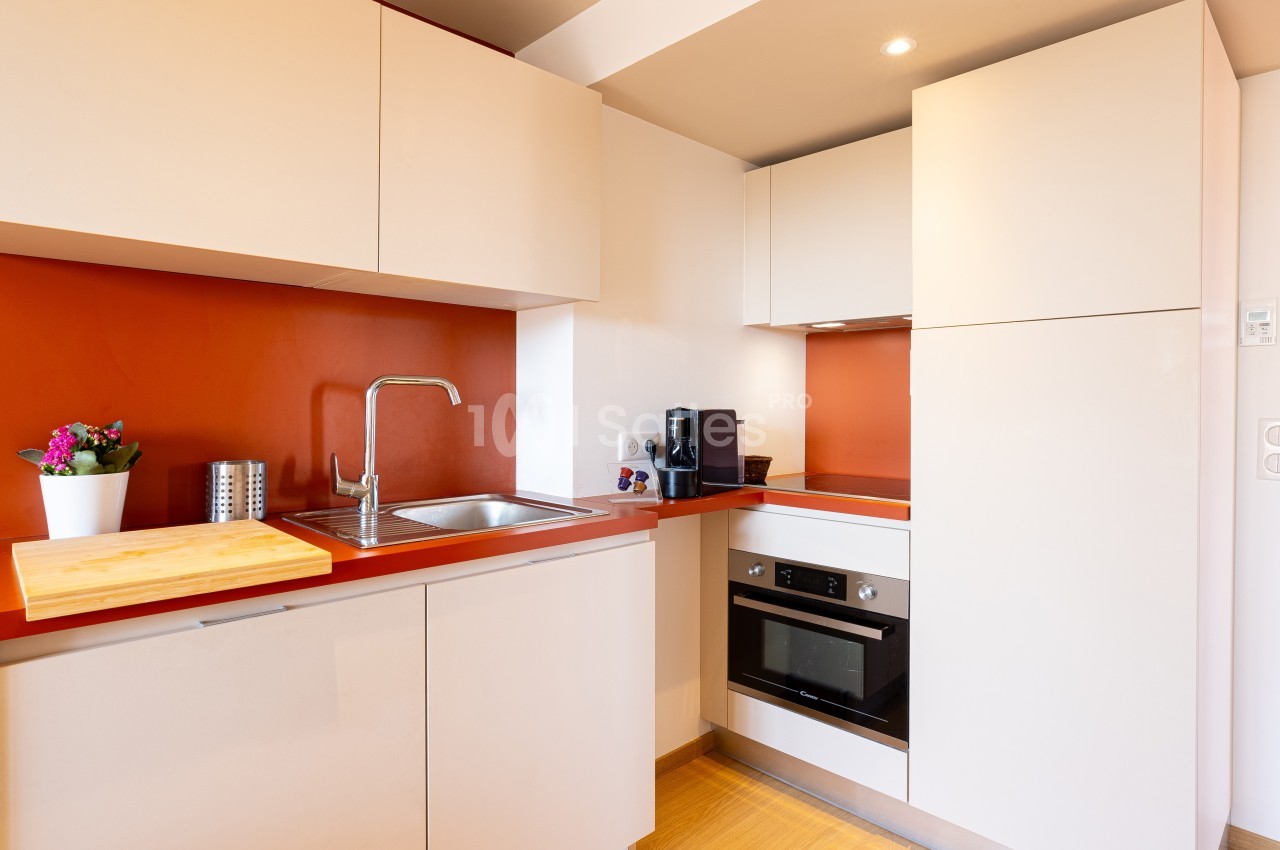 Cuisine moderne avec meubles beige, plan de travail rouge, évier inox, four encastré, et cafetière noire.