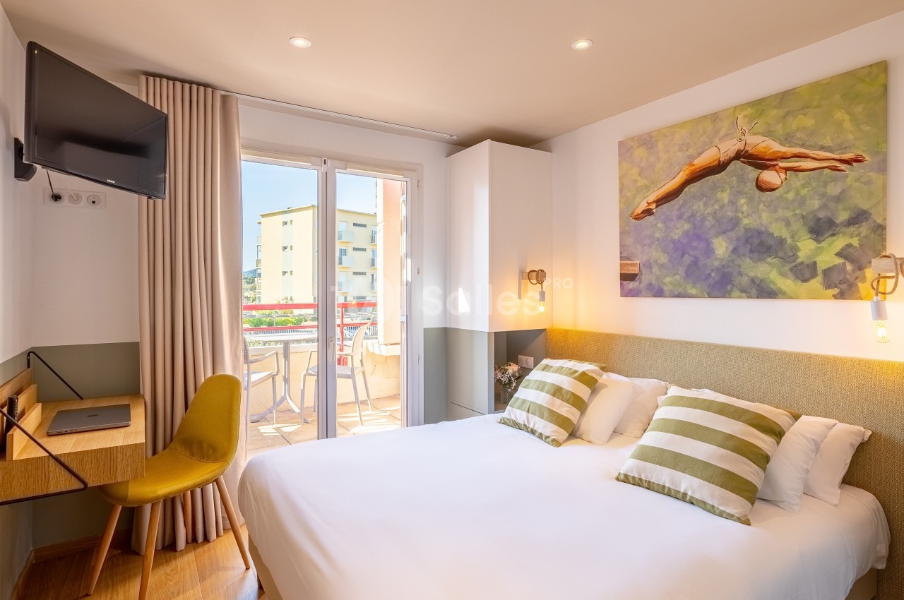 Chambre lumineuse avec lit double, bureau, télévision murale et accès à un balcon avec vue sur des bâtiments.