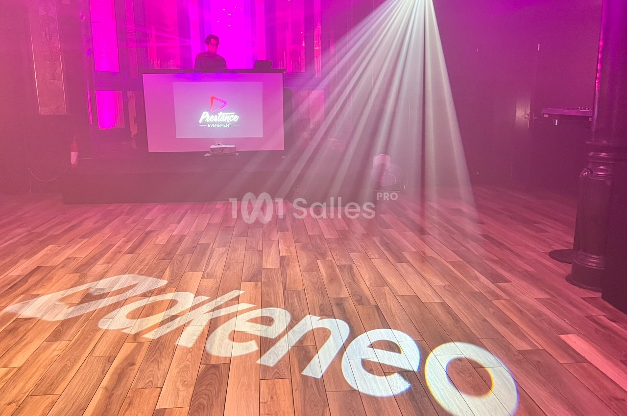 Salle de spectacle avec un DJ sur scène, éclairages colorés et logo ’Akeneo’ projeté sur le sol en bois.