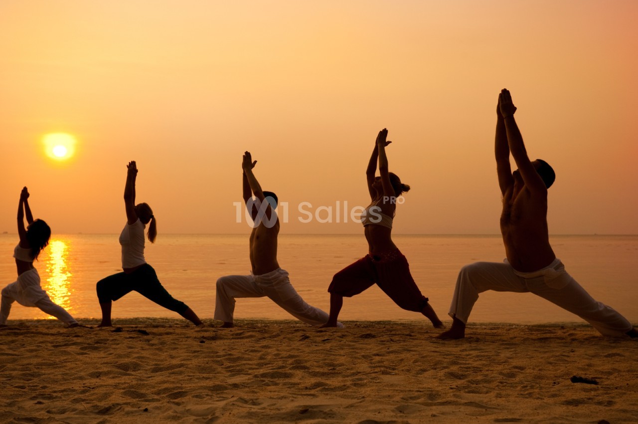 Personnes pratiquant le yoga sur une plage au coucher du soleil, en position debout avec les bras levés.