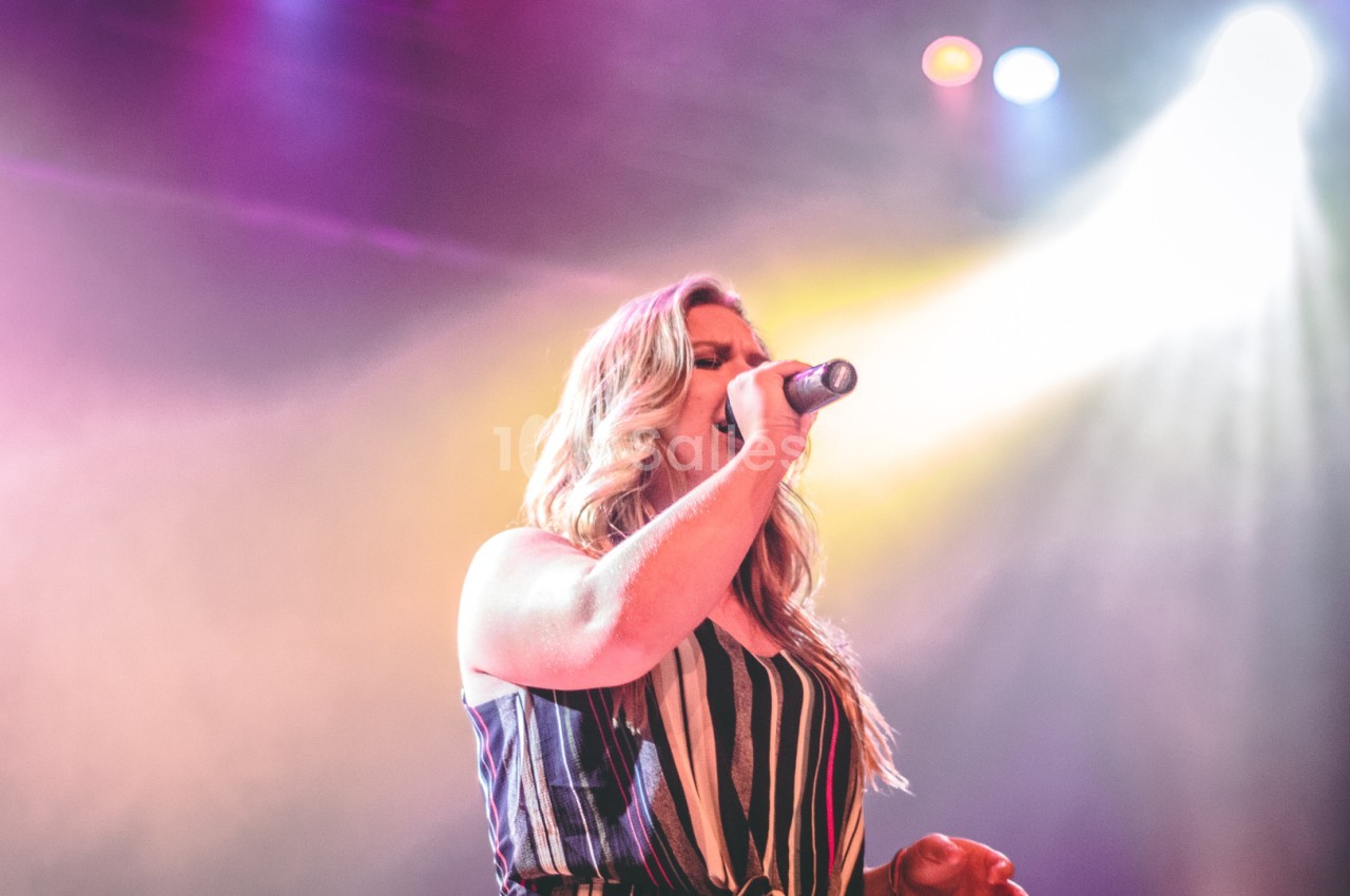 Une femme chante sur scène sous des projecteurs colorés, tenant un micro dans un environnement de concert.